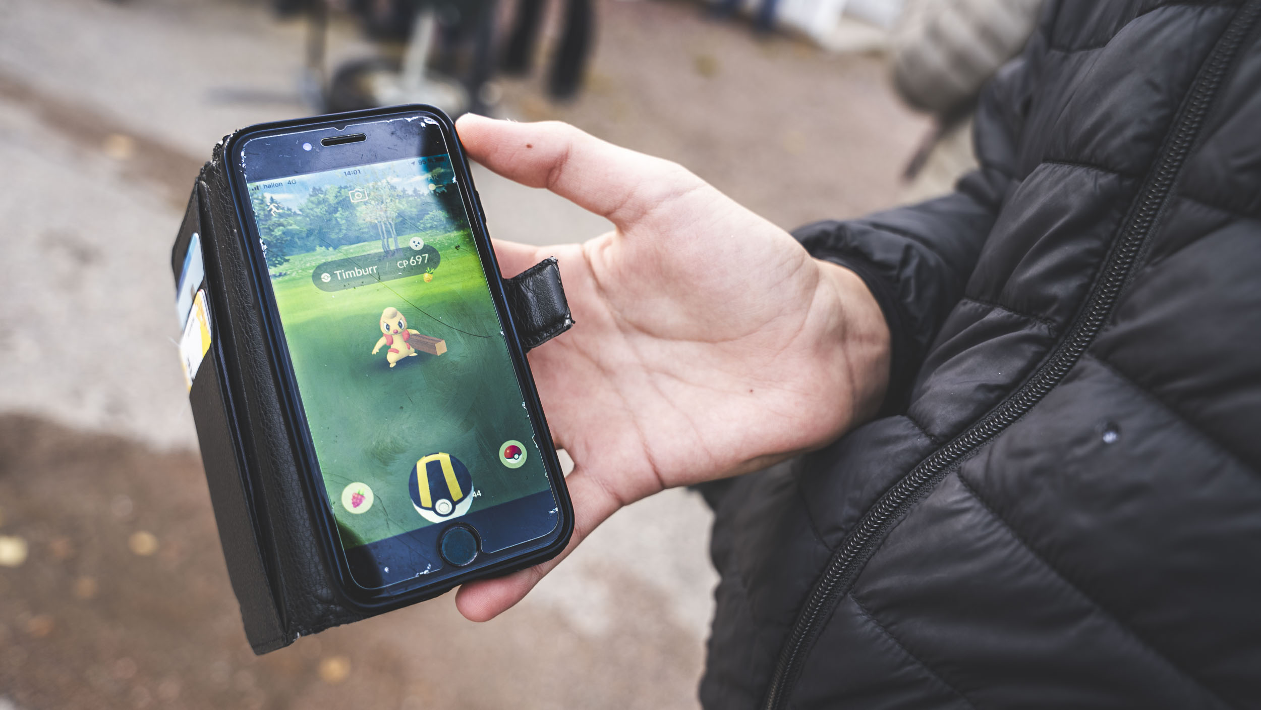 En person i vinterjacka håller i en mobiltelefon. På telefonens skärm är ett gult litet monster på en grön gräsmatta. Ovanför monstret är en textruta där det står Timburr CP697.