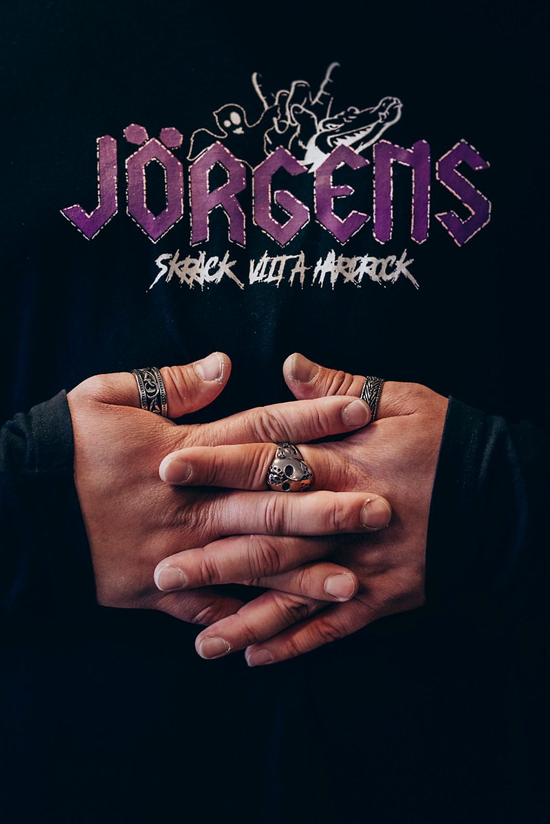 Ett par händer med silvriga dödskalleringar på fingrarna är knäppta över magen på en svart T-shirt med en logotyp på bröstet som säger "Jörgens Vilt, Skräck å Hårdrock".